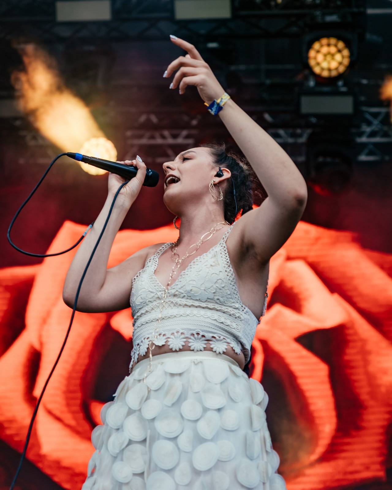 A female artist from the Revelland project holds the microphone and sings on stage. She is wearing a white top and skirt.