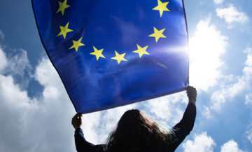 A woman holds the EU flag over her head in the wind. Clouds and the sky in the background.