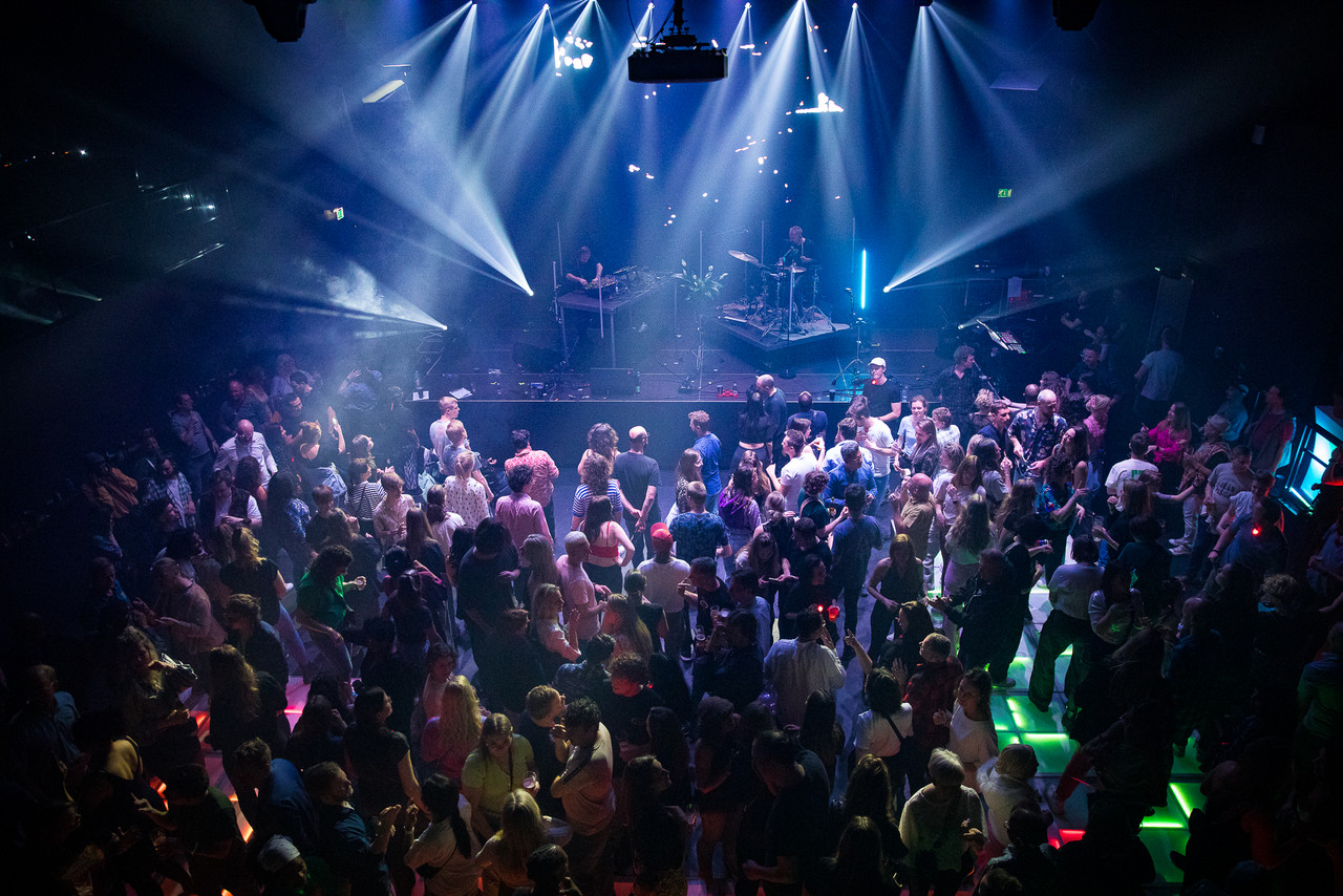 A birds eye view of a performance at Tivoli Vredenburg including the crowd and stage. There are strobe lights and the floor of the room has lights with different colours. 