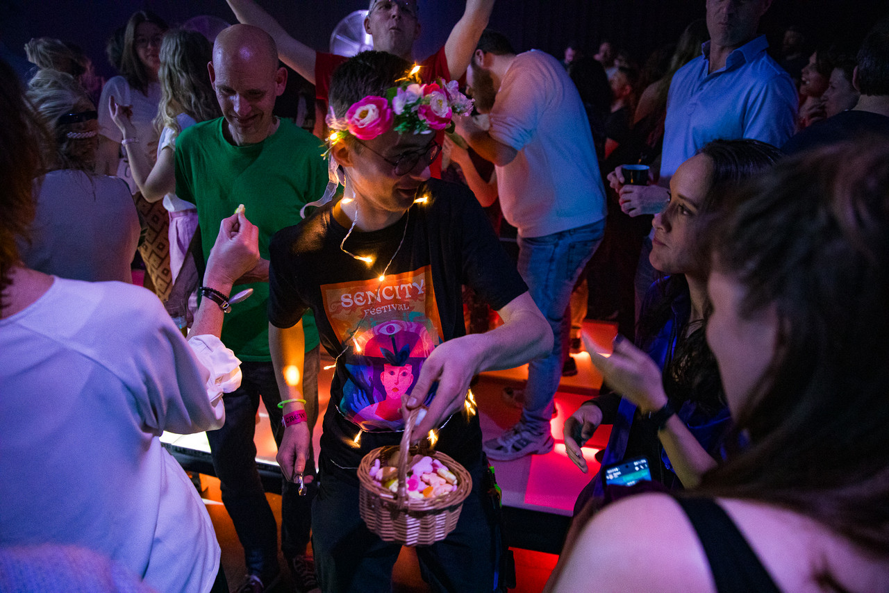 A young man holds a basket with sweets and has fairy lights around his neck and a flower crown on his head. He chats to a fellow female festival goer.