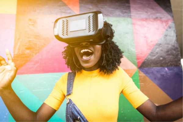 Image of a woman wearing a VR headset. She is wearing a yellow top, and smiling. Behind her is a multicoloured wall. 