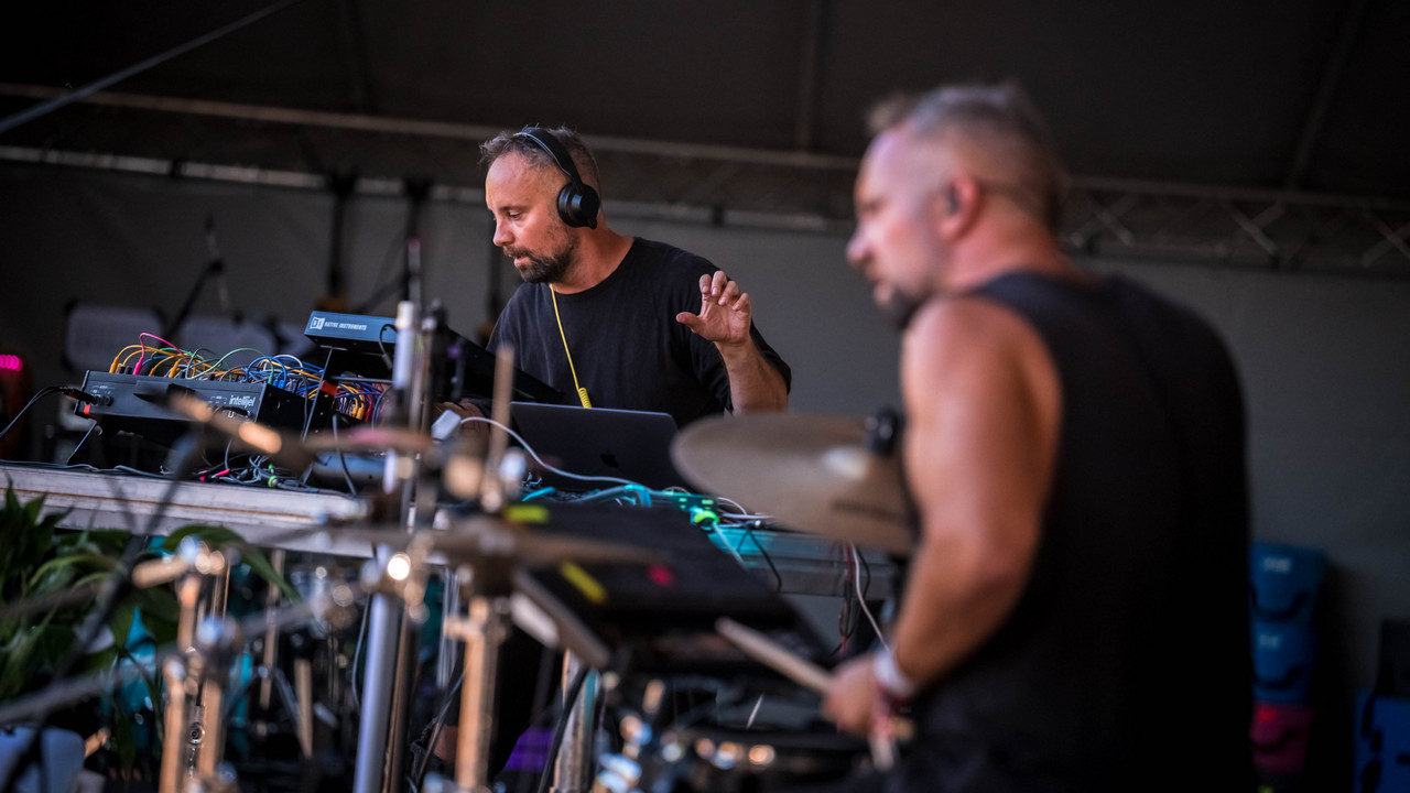 Two male musicians from the Revelland project perform on stage. One has headphones on and is on a DJ deck. The other man is playing the drums.