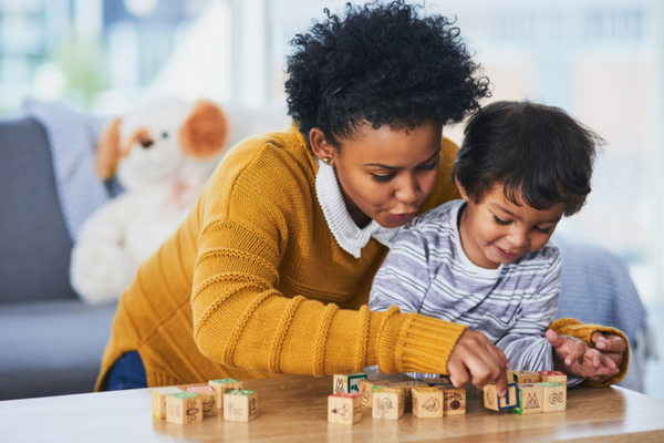 Image of a woman and a child, leading over a table and playing with building blocks. She is wearing a mustard coloured sweater and they are both smiling. 