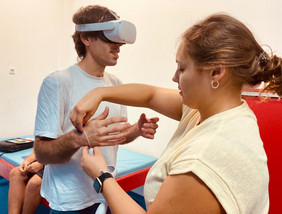 Image from the VIRTUS project in Portugal. A young man is wearing a VR set, and a woman is tying one of the controllers to his wrist. 