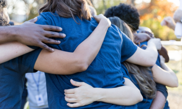 Image of a group of people hugging in a circle, showing their backs and arms around each other. They are all wearing blue t shirts. 