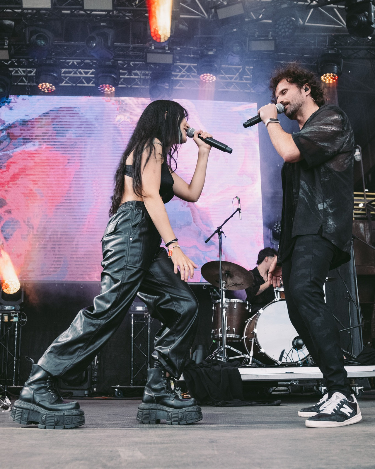 A female and male singer from the band K!NGDOM face each other on stage and sing into their microphones. She is wearing a black top, leather trousers and black chunky boots and the male singer is wearing a black shirt and trousers.