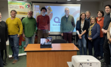 Image of the QoLAB partners all together in a room, with a table in the middle. On top of the table is a laptop showing another colleague attending online. 