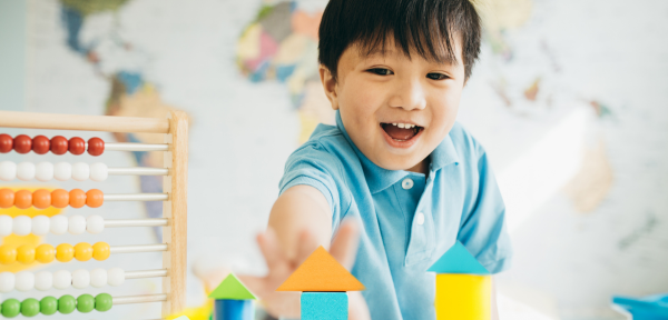Image of a young child wearing a blue shirt, playing with wooden toys on a table. He is smiling, and the room is sunny. 
