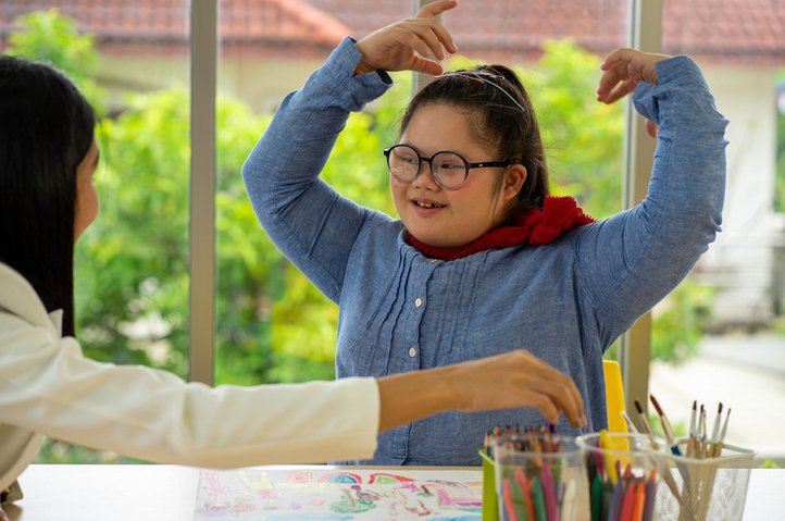 Image of a young girl with glasses, and her hair in a ponytail. She is smiling, and has her hands raised above her head. In front of her is a table with art supplies. 