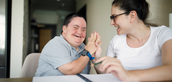 On the left of the visual is a woman with disabilities smiling as she holds the hand of a woman sitting next to her. The woman on the left is wearing a watch with a grey face and cobalt blue strap and a grey polo shirt, and has short brown hair. The woman on the right is smiling at the other woman, and wears black glasses and a white T-shirt. She has long brown hair that is tied up into a bun. They are both sitting at a wooden desk that has an opened blank notebook on it.