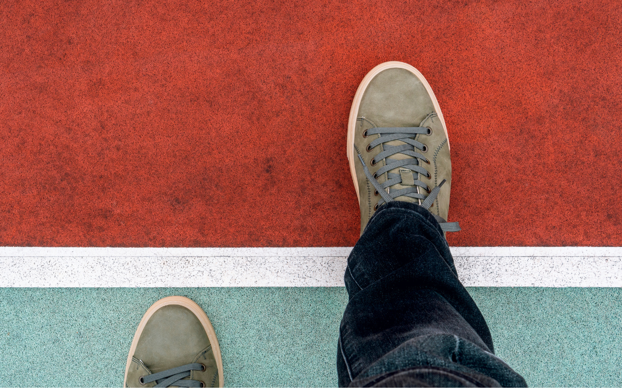 The image shows a floor that is red on the top half and turquoise on the bottom half. The two colours on the floor are separated by a white line. Two kaki trainers are positioned so that the left foot is on the bottom, turquoise part of the floor, and the right foot is stepping onto the red, upper portion of the floor.