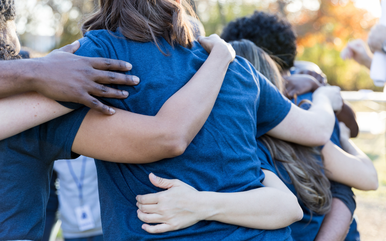 Image of a group of people hugging in a circle, showing their backs and arms around each other. They are all wearing blue t shirts. 