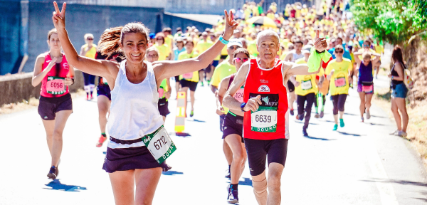 In the foreground, a woman and an old man are running. They smile and make the sign of victory. In the background, more people are running the marathon.