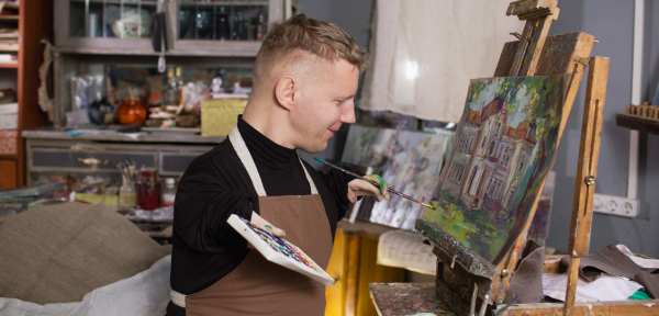 Image of a person with a physical disability paiting on a canvas. He is wearing a black t shirt, and has short blonde hair. He is smiling. 