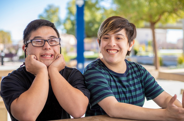 Two young people sitting at a table, in a park. One is wearing glasses, and one a blue striped shirt. They're both smiling. 