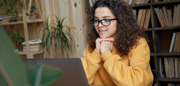 Image of a young girl with curly hear and glasses sitting in front of a laptop. She has her hands clasped together under her chin, smiling, and is wearing a yellow sweater. 